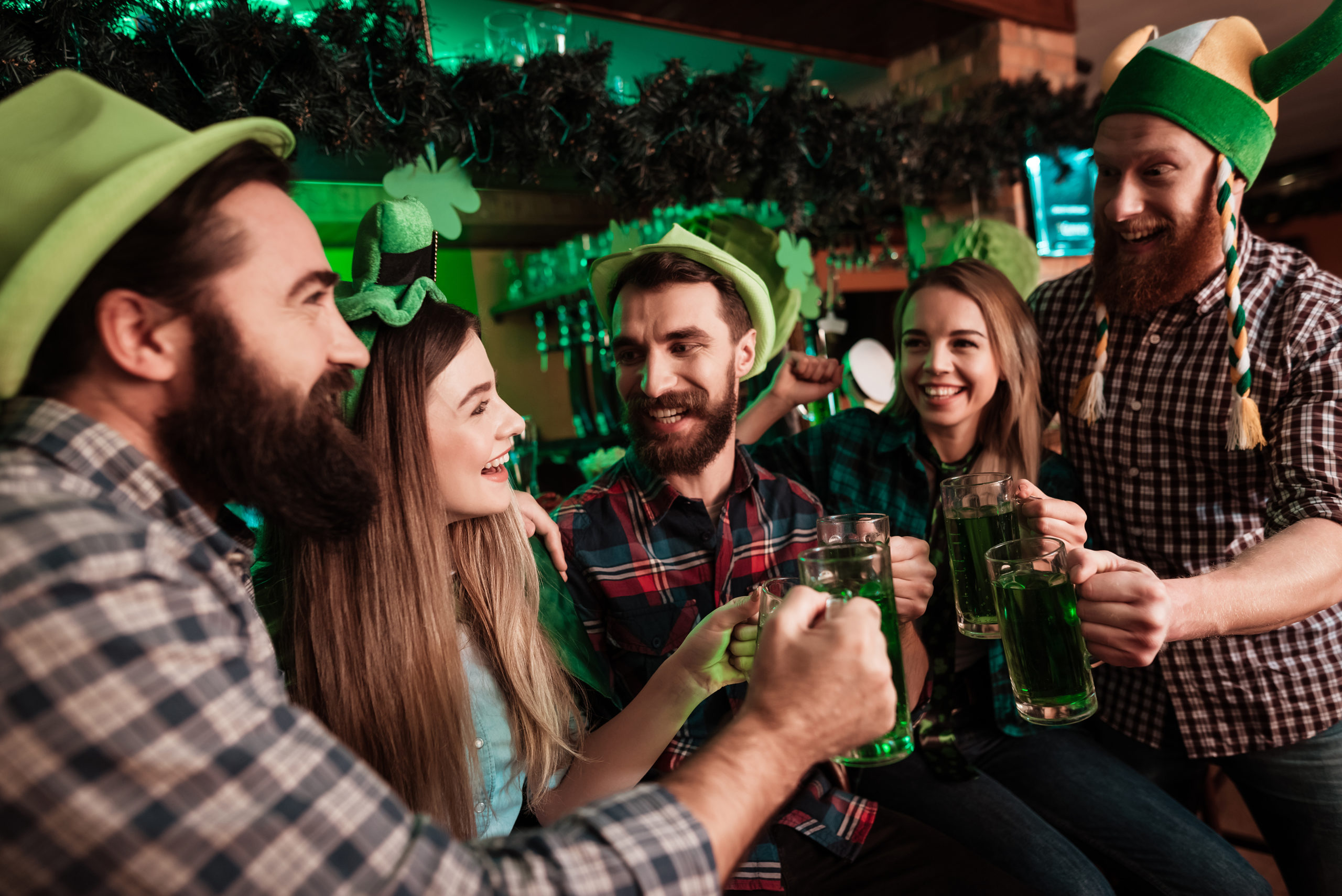 The company of young people celebrate St. Patrick's Day. They have fun at the bar. They are dressed in carnival headgear.